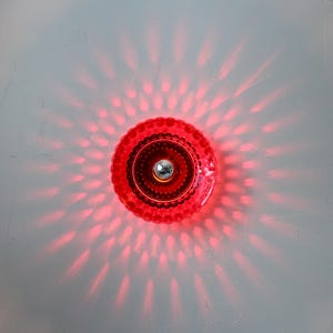K&ouml;nnte beinhalten: Eine rote, runde Leuchte mit strukturiertem, gewelltem Rand und einem silberfarbenen Mittelelement. Die Leuchte strahlt ein Muster roter Lichtstrahlen aus und erzeugt einen Sternausbruch-Effekt. Sie ist auf einer wei&szlig;en Oberfl&auml;che montiert.