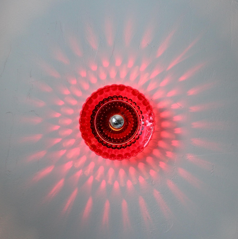 K&ouml;nnte beinhalten: Eine rote, runde Leuchte mit strukturiertem, gewelltem Rand und einem silberfarbenen Mittelelement. Die Leuchte strahlt ein Muster roter Lichtstrahlen aus und erzeugt einen Sternausbruch-Effekt. Sie ist auf einer wei&szlig;en Oberfl&auml;che montiert.