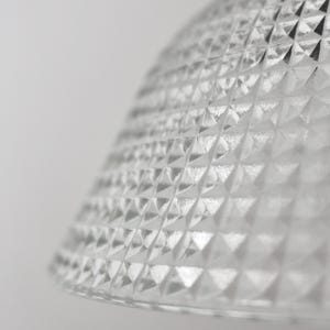 May include: Close-up of a clear glass lampshade with a diamond pattern. The lampshade has a rounded shape and a textured surface. The background is a plain white.