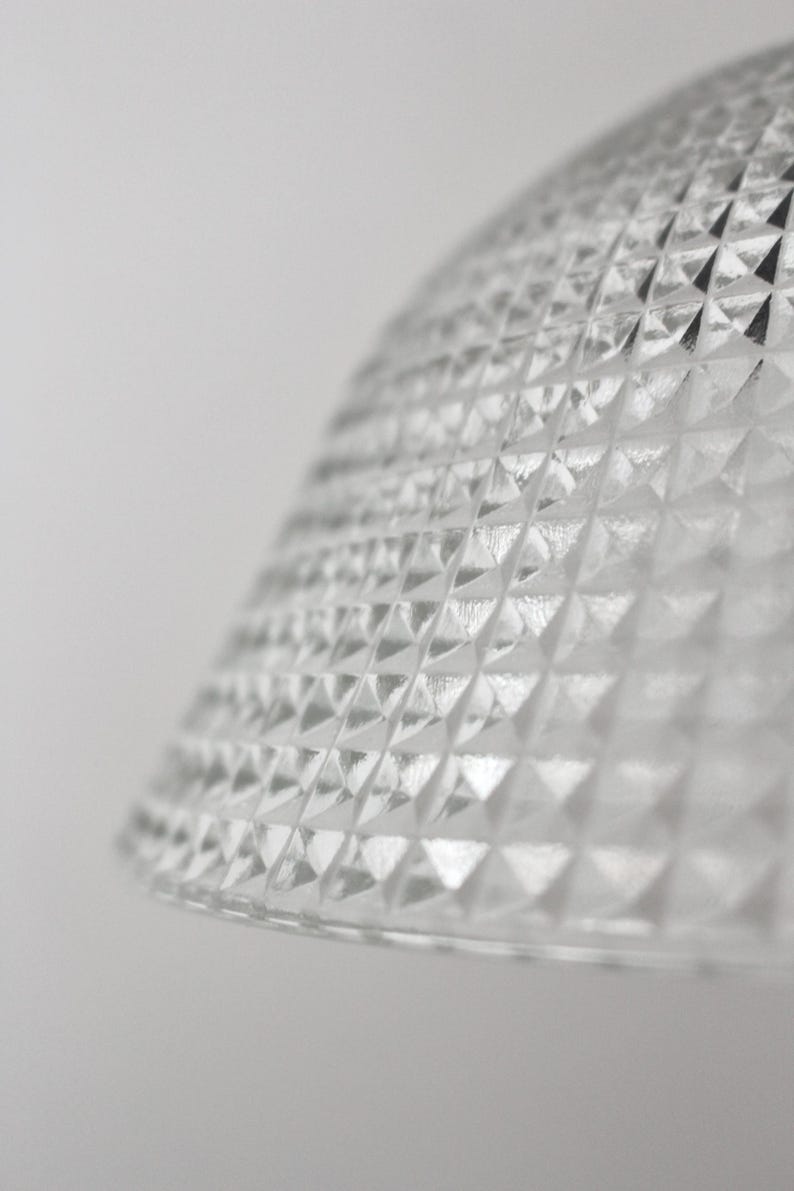 May include: Close-up of a clear glass lampshade with a diamond pattern. The lampshade has a rounded shape and a textured surface. The background is a plain white.