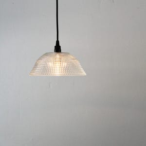 May include: Clear glass pendant light with a textured, geometric pattern. The light fixture has a conical shade and a black cord. The light is illuminated, casting a warm glow. The background is a plain white wall.