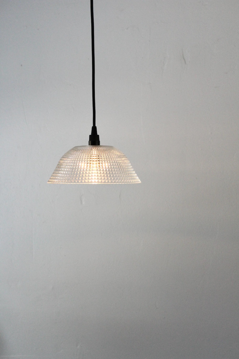 May include: Clear glass pendant light with a textured, geometric pattern. The light fixture has a conical shade and a black cord. The light is illuminated, casting a warm glow. The background is a plain white wall.
