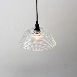 May include: Clear glass pendant light with a textured, geometric pattern on the shade. The light fixture hangs from a black cord and a black fixture. The light is on, illuminating the shade.