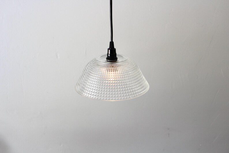 May include: Clear glass pendant light with a textured, geometric pattern on the shade. The light fixture hangs from a black cord and a black fixture. The light is on, illuminating the shade.