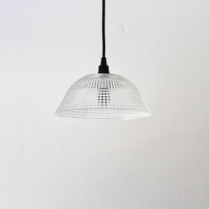 May include: Clear glass pendant light with a textured, geometric pattern. The light fixture hangs from a black cord and canopy against a white background. The shade is dome-shaped.