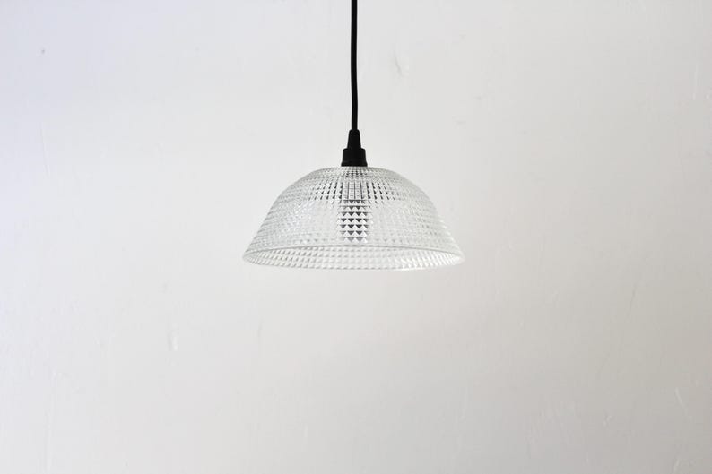 May include: Clear glass pendant light with a textured, geometric pattern. The light fixture hangs from a black cord and canopy against a white background. The shade is dome-shaped.