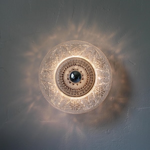 May include: A clear glass ceiling light fixture with an ornate floral design. The light has a central, round, silver-colored element and is surrounded by a decorative, etched pattern. The light casts a warm glow.