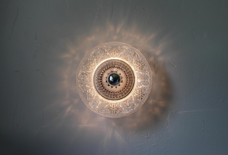 May include: A clear glass ceiling light fixture with an ornate floral design. The light has a central, round, silver-colored element and is surrounded by a decorative, etched pattern. The light casts a warm glow.