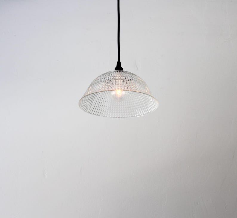 May include: Clear glass pendant light with a textured, dotted pattern. The light fixture has a bell-shaped shade and a black cord. The light bulb is visible inside the shade, illuminating the space. The background is a plain white wall.