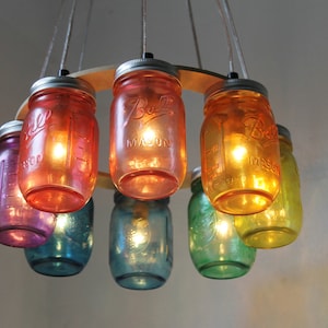 May include: A rainbow-colored chandelier made from mason jars with light bulbs inside. The jars are hung from a wooden ring.