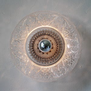 May include: A clear glass light fixture with a floral pattern around the edge. The center features a silver orb and a textured, circular design. The fixture is mounted on a white wall, creating a decorative lighting element.