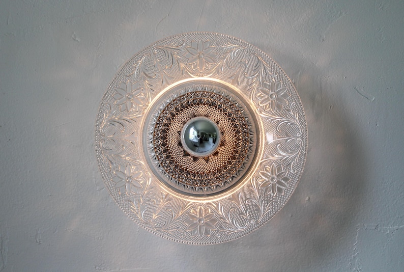 May include: A clear glass light fixture with a floral pattern around the edge. The center features a silver orb and a textured, circular design. The fixture is mounted on a white wall, creating a decorative lighting element.