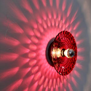 K&ouml;nnte beinhalten: Rote Wandleuchte aus Glas mit strukturiertem Design. Das Licht wirft ein Muster aus rotem Licht auf die Wand und erzeugt einen einzigartigen visuellen Effekt. Die Leuchte hat eine Vintage-&Auml;sthetik. Ma&szlig;e in cm.