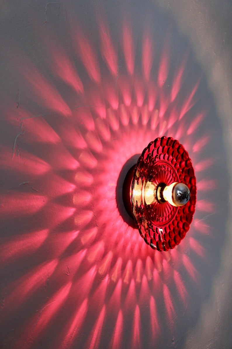 K&ouml;nnte beinhalten: Rote Wandleuchte aus Glas mit strukturiertem Design. Das Licht wirft ein Muster aus rotem Licht auf die Wand und erzeugt einen einzigartigen visuellen Effekt. Die Leuchte hat eine Vintage-&Auml;sthetik. Ma&szlig;e in cm.