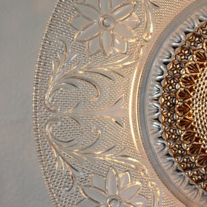 May include: Close-up of a clear glass plate with intricate floral and leaf patterns. The plate features embossed flowers and a textured background, with a detailed center design. The plate is a decorative piece.