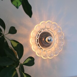 May include: A clear glass wall sconce with a scalloped edge and a round, frosted glass center. The sconce has a single light bulb in the center.