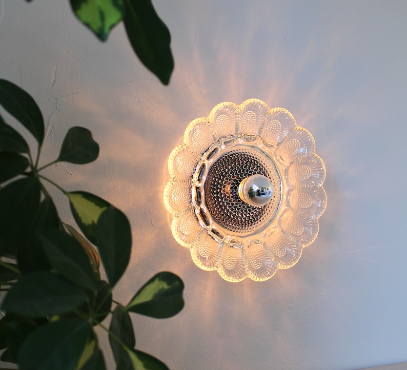 May include: A clear glass wall sconce with a scalloped edge and a round, frosted glass center. The sconce has a single light bulb in the center.