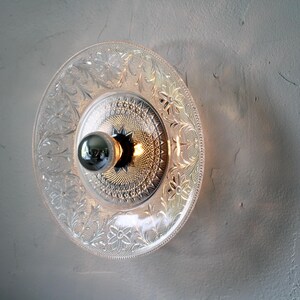 May include: A clear glass plate light fixture with a decorative floral pattern. The light fixture has a silver bulb and a silver center. The light is mounted on a light gray wall.