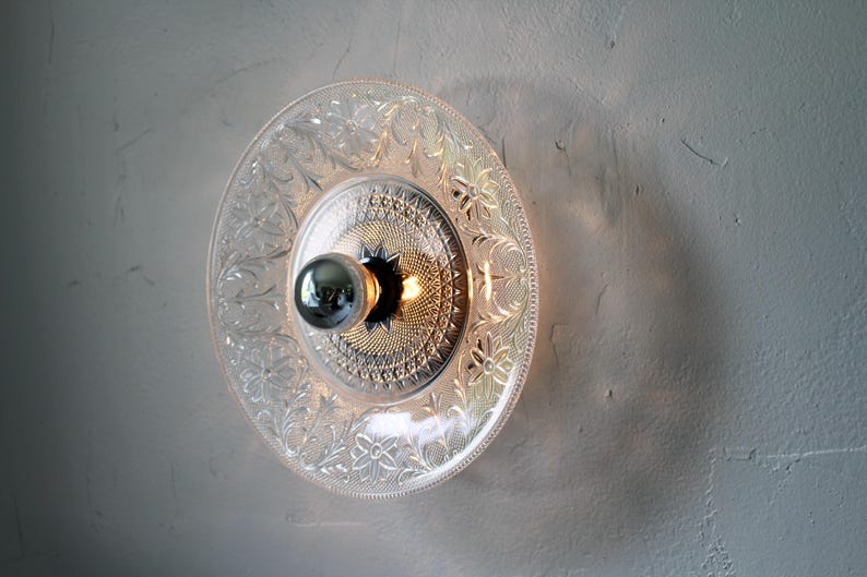May include: A clear glass plate light fixture with a decorative floral pattern. The light fixture has a silver bulb and a silver center. The light is mounted on a light gray wall.