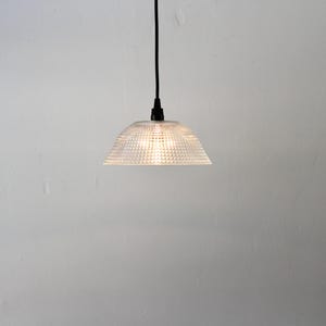 May include: A clear, textured glass pendant light with a dome-shaped shade. The light hangs from a black cord and is illuminated, casting a warm glow. The fixture is a modern design, suitable for interior lighting.