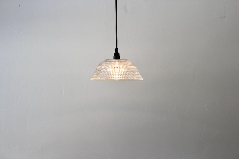May include: A clear, textured glass pendant light with a dome-shaped shade. The light hangs from a black cord and is illuminated, casting a warm glow. The fixture is a modern design, suitable for interior lighting.