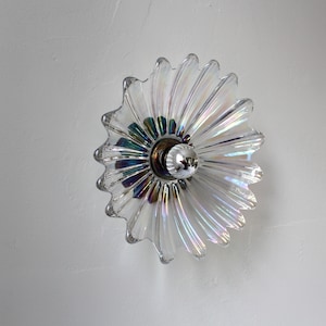 May include: A clear glass ceiling light fixture with a iridescent finish in the shape of a flower. The light fixture has a silver metal base and a single light bulb.