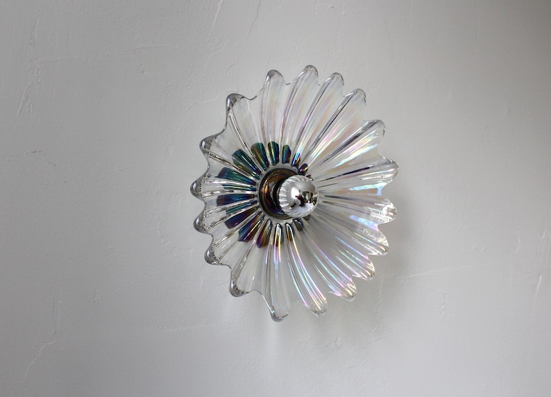 May include: A clear glass ceiling light fixture with a iridescent finish in the shape of a flower. The light fixture has a silver metal base and a single light bulb.