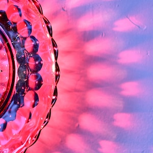 K&ouml;nnte beinhalten: Nahaufnahme eines durchsichtigen, dekorativen Glas tellers mit gewelltem Rand. Der Teller wird von rotem Licht beleuchtet, das herzf&ouml;rmige Schatten auf eine violett-blaue Oberfl&auml;che wirft.
