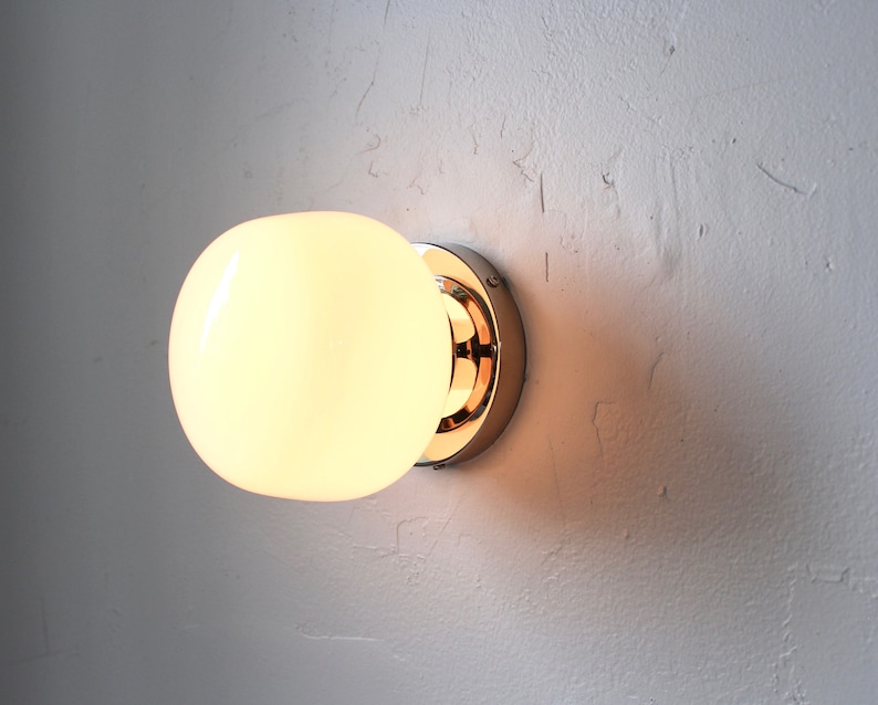 Op de afbeelding: Een chromen wandlamp met een witte, matte glazen bol. Het licht is aan en de bol is verlicht.