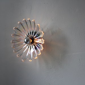May include: A clear glass wall sconce with a flower-shaped design. The sconce has a light bulb in the center and casts a warm glow.