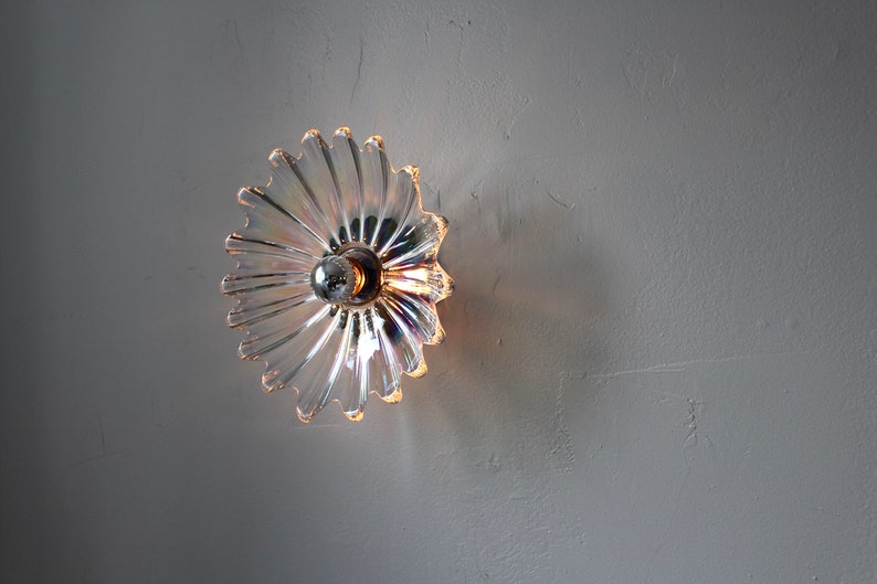 May include: A clear glass wall sconce with a flower-shaped design. The sconce has a light bulb in the center and casts a warm glow.