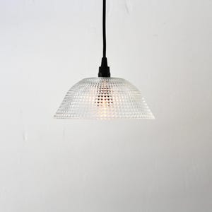 May include: Clear glass pendant light with a textured, geometric pattern. The light fixture has a dome shape and hangs from a black cord. The light is turned on, illuminating the interior.