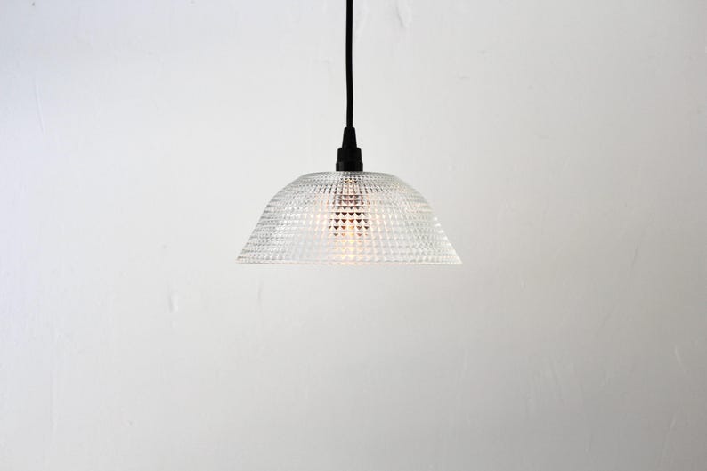 May include: Clear glass pendant light with a textured, geometric pattern. The light fixture has a dome shape and hangs from a black cord. The light is turned on, illuminating the interior.