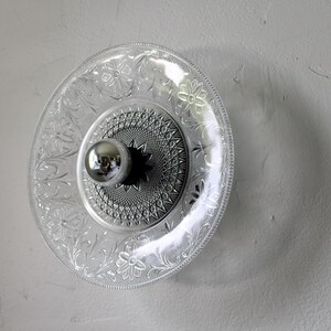 May include: A clear glass plate with a floral and geometric pattern, mounted on a wall. A silver-colored light fixture is centered on the plate. The plate is approximately 10 inches in diameter.