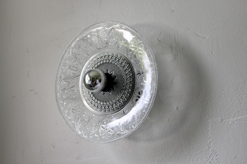 May include: A clear glass plate with a floral and geometric pattern, mounted on a wall. A silver-colored light fixture is centered on the plate. The plate is approximately 10 inches in diameter.