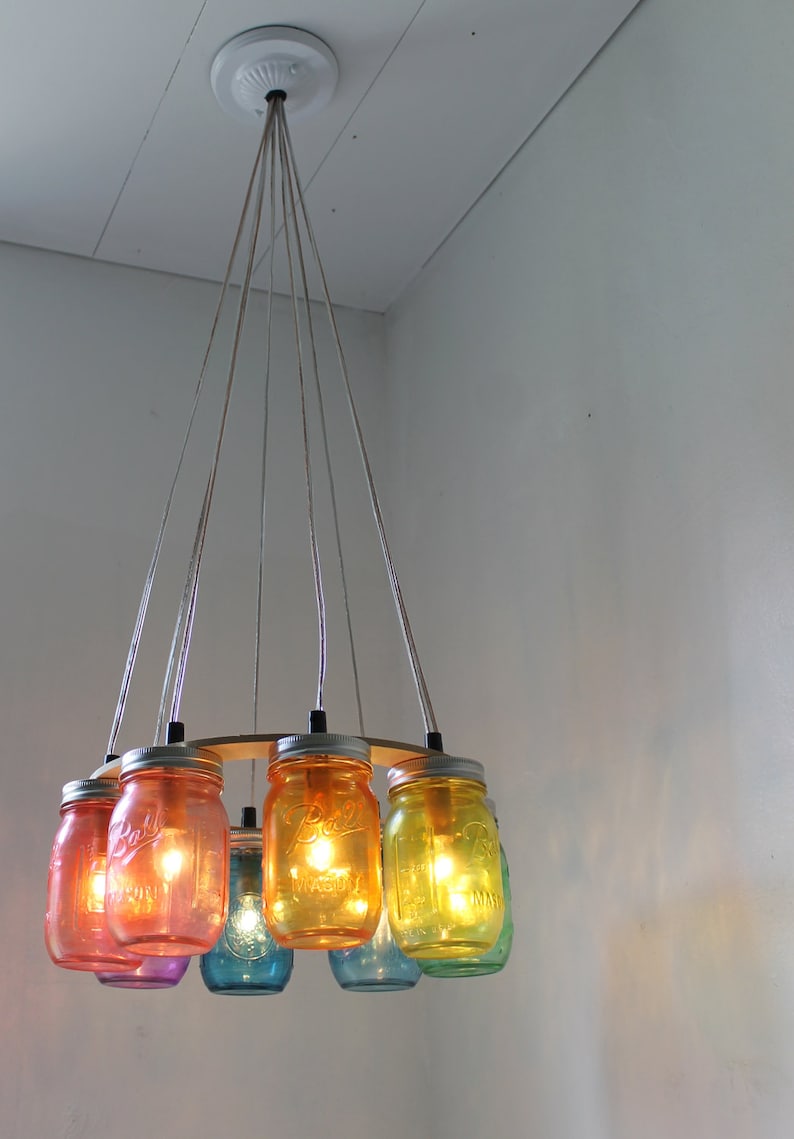 May include: A rainbow colored glass jar chandelier with eight jars hanging from a circular wooden base. The jars are lit with light bulbs and have a vintage look.
