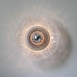 May include: A clear glass ceiling light fixture with a decorative floral pattern around the edge. The center features a textured design and a silver-colored, spherical element. The light is illuminated, creating a warm glow.