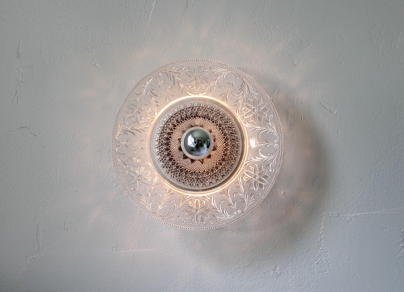 May include: A clear glass ceiling light fixture with a decorative floral pattern around the edge. The center features a textured design and a silver-colored, spherical element. The light is illuminated, creating a warm glow.