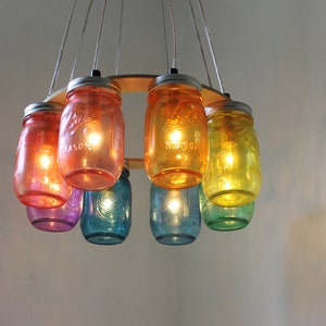 May include: A rainbow-colored glass jar chandelier with seven jars hanging from a wooden ring. The jars are lit with light bulbs and have a warm glow.
