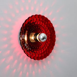 K&ouml;nnte beinhalten: Rote Wandleuchte aus Glas mit strukturiertem, kreisf&ouml;rmigem Schirm. Die Leuchte hat eine silberne Gl&uuml;hbirne und wirft einen roten Schein an die Wand. Der Schirm hat ein dekoratives, blasenartiges Muster.