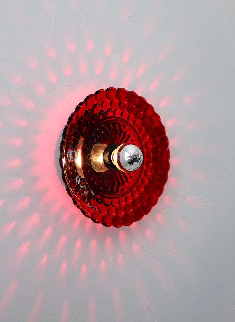 K&ouml;nnte beinhalten: Rote Wandleuchte aus Glas mit strukturiertem, kreisf&ouml;rmigem Schirm. Die Leuchte hat eine silberne Gl&uuml;hbirne und wirft einen roten Schein an die Wand. Der Schirm hat ein dekoratives, blasenartiges Muster.