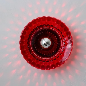 K&ouml;nnte beinhalten: Rote Wandleuchte mit strukturiertem, gewelltem Rand. In der Mitte befindet sich eine silberne Kugel. Rotes Licht geht von der Leuchte aus und erzeugt ein Muster an der wei&szlig;en Wand. Dekoratives Leuchtmittel.