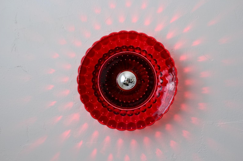 K&ouml;nnte beinhalten: Rote Wandleuchte mit strukturiertem, gewelltem Rand. In der Mitte befindet sich eine silberne Kugel. Rotes Licht geht von der Leuchte aus und erzeugt ein Muster an der wei&szlig;en Wand. Dekoratives Leuchtmittel.