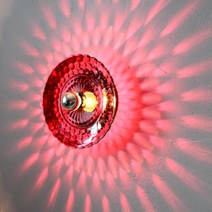K&ouml;nnte beinhalten: Eine rote Glas-Wandleuchte mit strukturiertem, gewelltem Rand. Das Licht wirft ein radiales Muster aus rotem Licht auf die Wand. Die Leuchte hat eine silberfarbene Gl&uuml;hbirnenfassung und eine warmtonige Gl&uuml;hbirne.