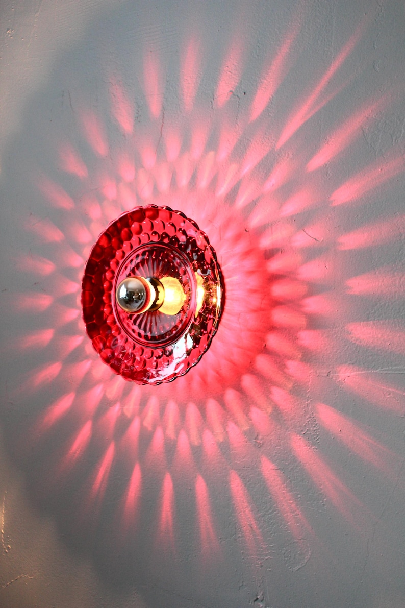 K&ouml;nnte beinhalten: Eine rote Glas-Wandleuchte mit strukturiertem, gewelltem Rand. Das Licht wirft ein radiales Muster aus rotem Licht auf die Wand. Die Leuchte hat eine silberfarbene Gl&uuml;hbirnenfassung und eine warmtonige Gl&uuml;hbirne.