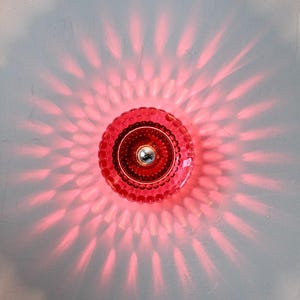 K&ouml;nnte beinhalten: Eine rote Glasleuchte mit einem strukturierten, kreisf&ouml;rmigen Design. Das Licht wirft ein radiales Muster aus rosa Licht auf die Wand. Die Leuchte hat eine silberfarbene Mitte.