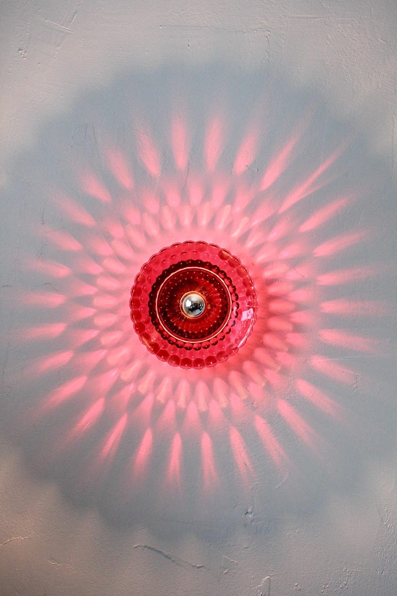 K&ouml;nnte beinhalten: Eine rote Glasleuchte mit einem strukturierten, kreisf&ouml;rmigen Design. Das Licht wirft ein radiales Muster aus rosa Licht auf die Wand. Die Leuchte hat eine silberfarbene Mitte.