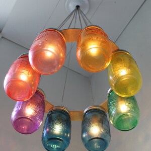 May include: A circular chandelier made of repurposed glass jars painted in various colors, including purple, blue, green, orange, and yellow. The jars are suspended from a wooden ring and have light bulbs inside.
