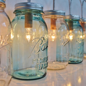 May include: A row of mason jars with light bulbs inside, hanging from the ceiling. The jars are clear glass with a blue tint and have a silver lid. The light bulbs are lit and casting a warm glow.
