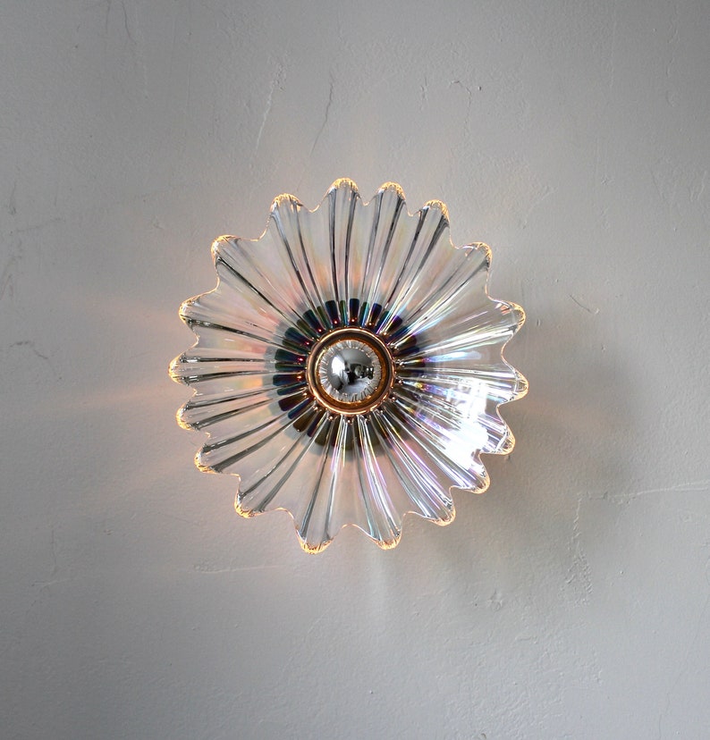 May include: A clear iridescent glass light fixture shaped like a flower with a round center. The fixture has a white background and is mounted on a wall.